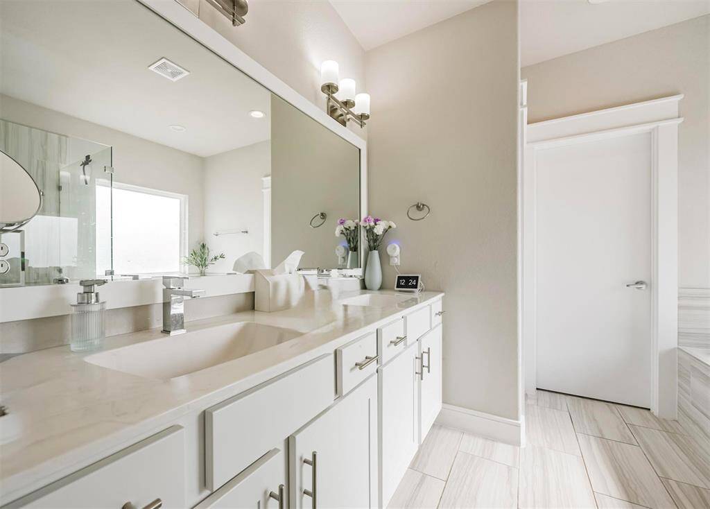 Primary bathroom featuring a double vanity with ample storage, a large mirror, and tasteful lighting complimented by neutral tile flooring and a clean, contemporary design ideal for a variety of personal styles.