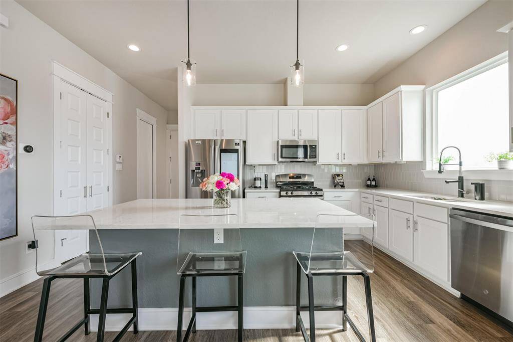 This modern, well-lit kitchen features sleek white cabinetry, stainless steel appliances, a large island with seating, quartz countertops and sleek pendant lighting.