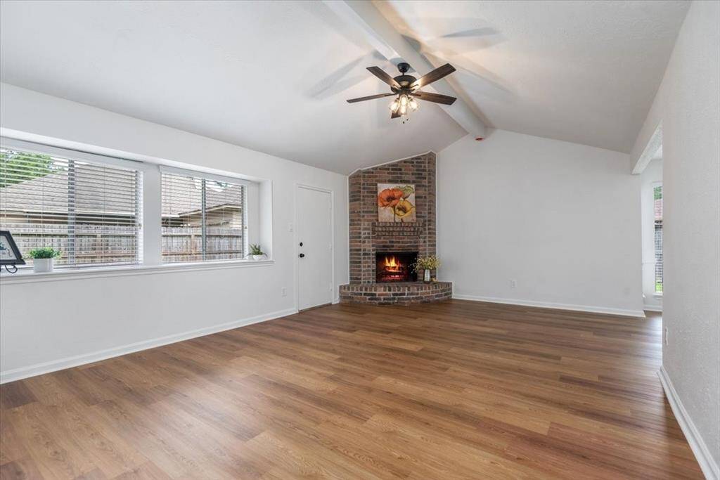 This spacious living room features a vaulted ceiling with a cozy brick fireplace, and abundant natural light from large windows, complemented by stylish luxury vinyl flooring.