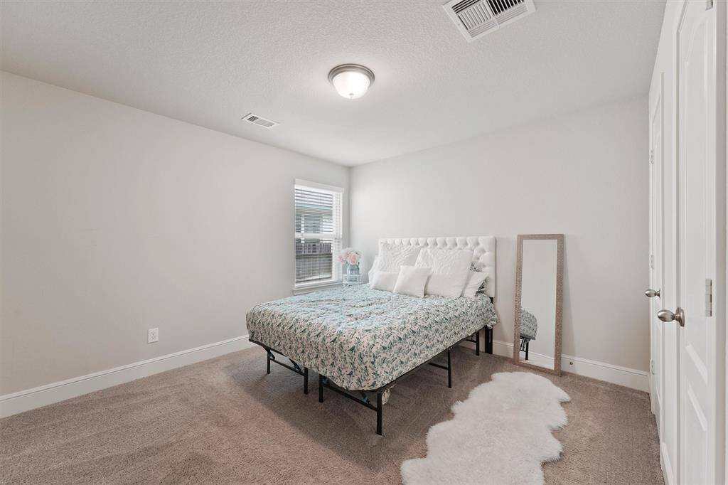 Cozy bedroom with neutral walls and carpet, featuring a comfortable bed with plush headboard. A large window offers natural light, and there's a stylish floor mirror and fluffy rug for added charm.