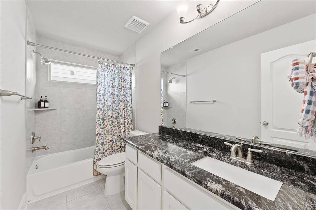 Modern bathroom with elegant black granite countertops and dual sinks. It features a shower-tub combo with a floral curtain, ample lighting, and neutral tile flooring.