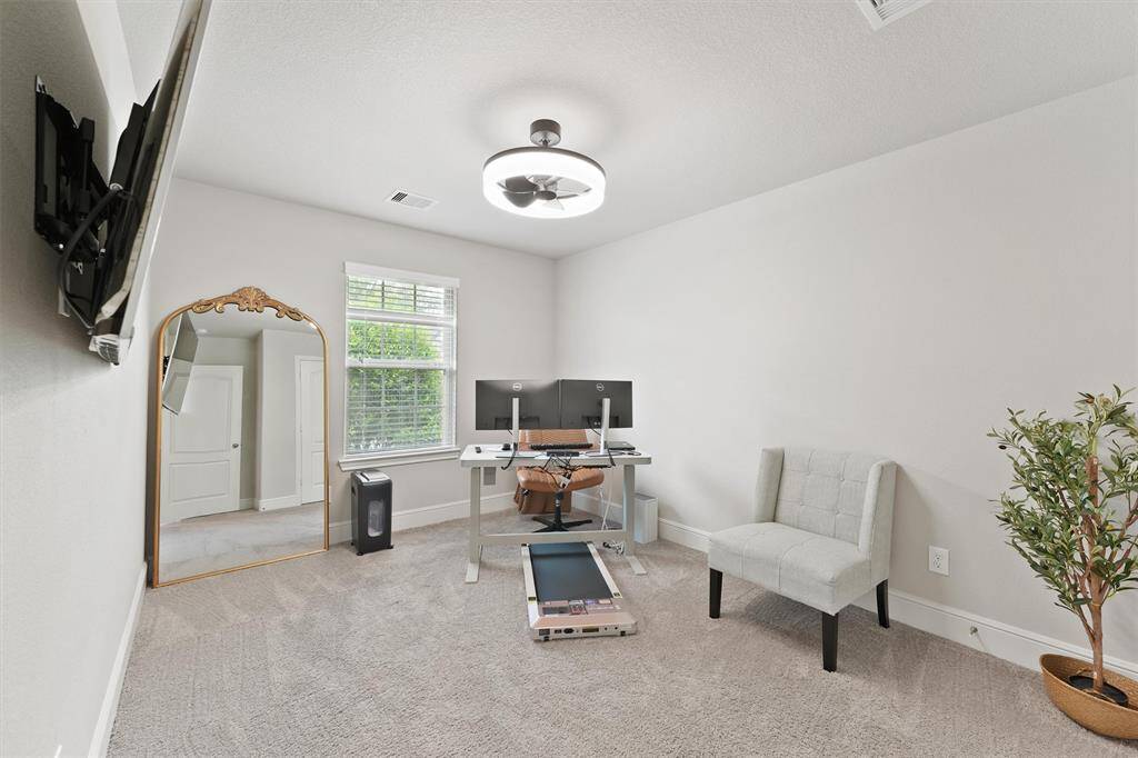 This room is a bright and versatile space ideal for a home office. It features a large window for natural light, a modern desk setup, a wall-mounted TV, and a comfortable chair. The neutral decor and carpeted floor create a cozy atmosphere.