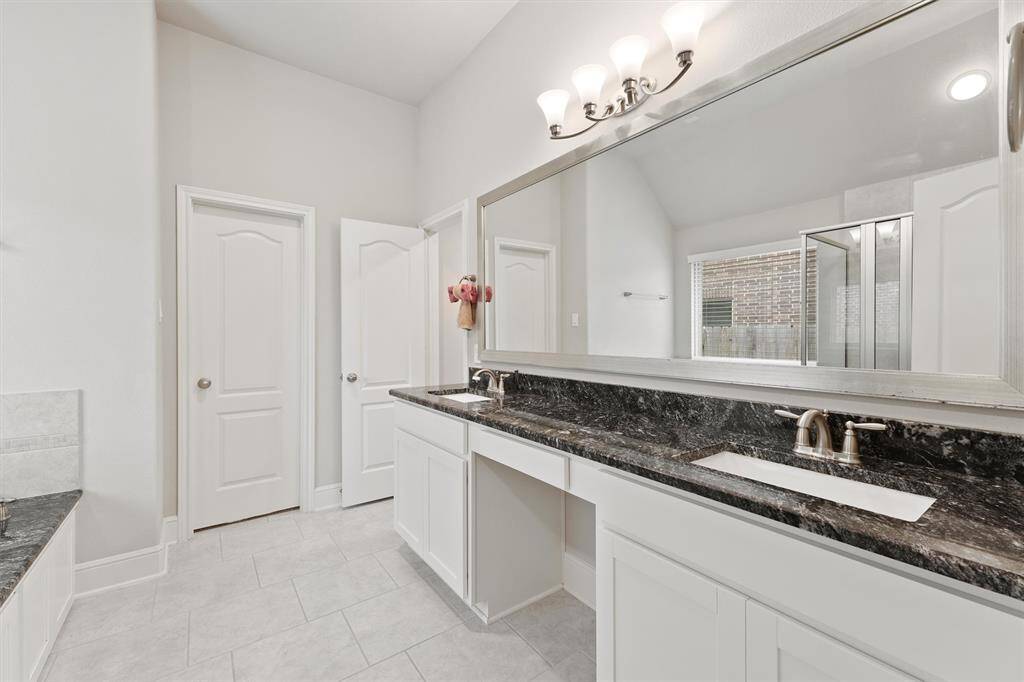 This bathroom features a spacious design with dual sinks, a large mirror, and elegant black granite countertops. It offers ample storage with white cabinets and has a separate shower area. The neutral color palette and natural light create a bright and inviting atmosphere.