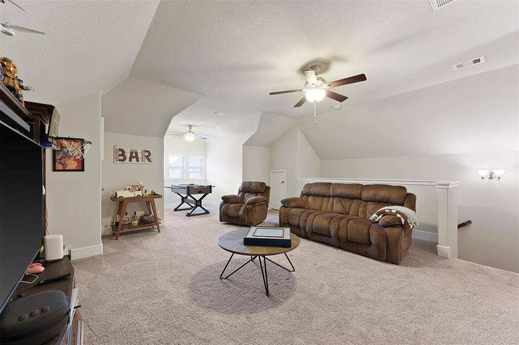 This spacious attic room is ideal for relaxation and entertainment. It features cozy seating, a ceiling fan, and a corner bar area. Natural light filters through a window, adding warmth to the carpeted space. Perfect for a game room or lounge.