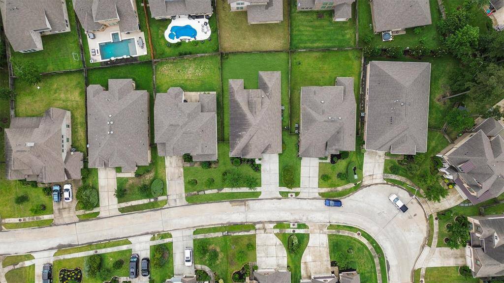This aerial photo shows a suburban neighborhood with well-maintained homes, each featuring neat lawns and driveways. One property has a backyard pool, and the area appears quiet and residential with tree-lined streets.