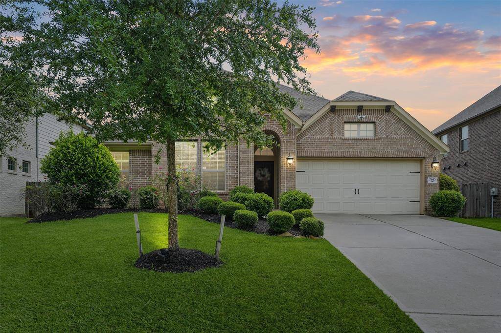 This inviting brick home features a well-maintained front yard with lush greenery and a mature tree. It has a two-car garage and a welcoming pathway leading to the entrance, enhanced by a beautiful sunset backdrop.