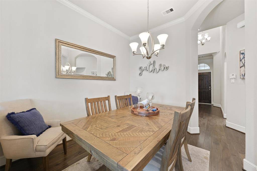 This dining area features a rustic wooden table with seating for six, a cozy armchair, and a decorative mirror. The walls are painted a soft neutral color, and a stylish chandelier adds warmth. A welcoming entryway with a dark wood door is visible in the background.