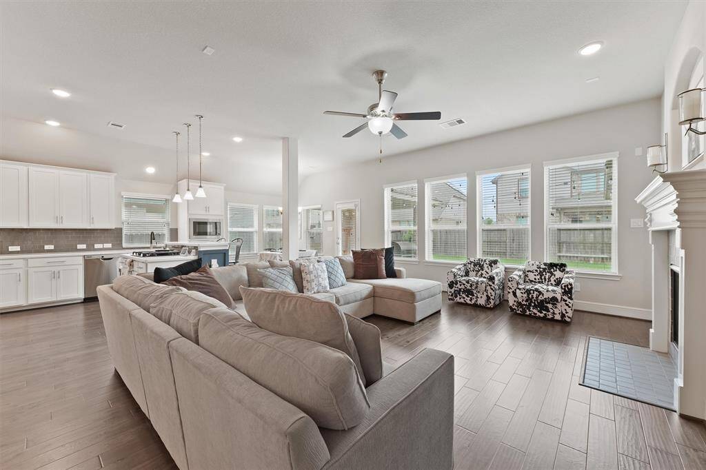 This spacious, open-concept living area features a modern kitchen with white cabinetry, stainless steel appliances, and a large island. The adjoining living room has a cozy sectional, large windows for natural light, and a fireplace, perfect for entertaining or relaxing.