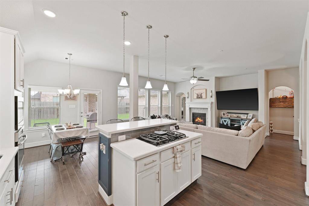 This photo shows a spacious open-concept kitchen and living area. The kitchen features a central island with a gas cooktop, white cabinetry, and modern pendant lighting. The adjacent dining area has large windows and a door leading to the backyard. The living room includes a cozy fireplace, ceiling fan, and a large TV, offering a comfortable and inviting space.