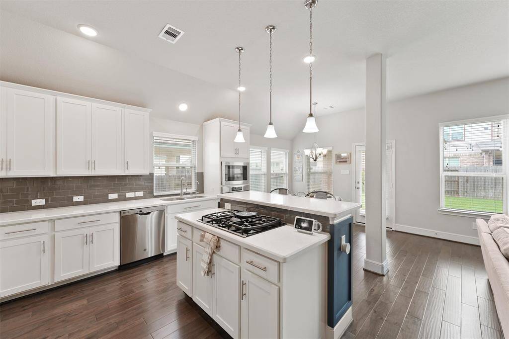 This photo showcases a modern kitchen with white cabinetry, stainless steel appliances, and a central island featuring a gas cooktop. The space is bright and airy, complemented by pendant lighting and dark hardwood floors. Large windows provide plenty of natural light and a view of the backyard.
