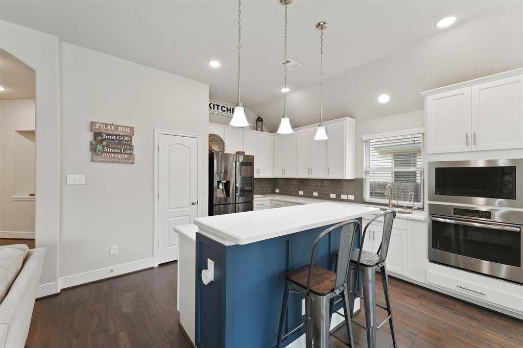 This kitchen features a modern design with white cabinetry, stainless steel appliances, and a central island with a blue accent and seating. Pendant lights and ample natural light enhance the space's bright, open feel.