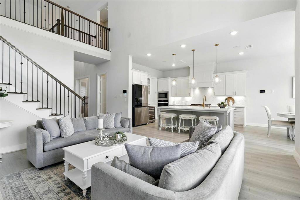 This open-concept living area features a modern kitchen with a large island, elegant pendant lighting, and stainless steel appliances. The space is bright and airy, with high ceilings, A staircase with a wrought iron railing leads to an upper level, enhancing the home's spacious feel.