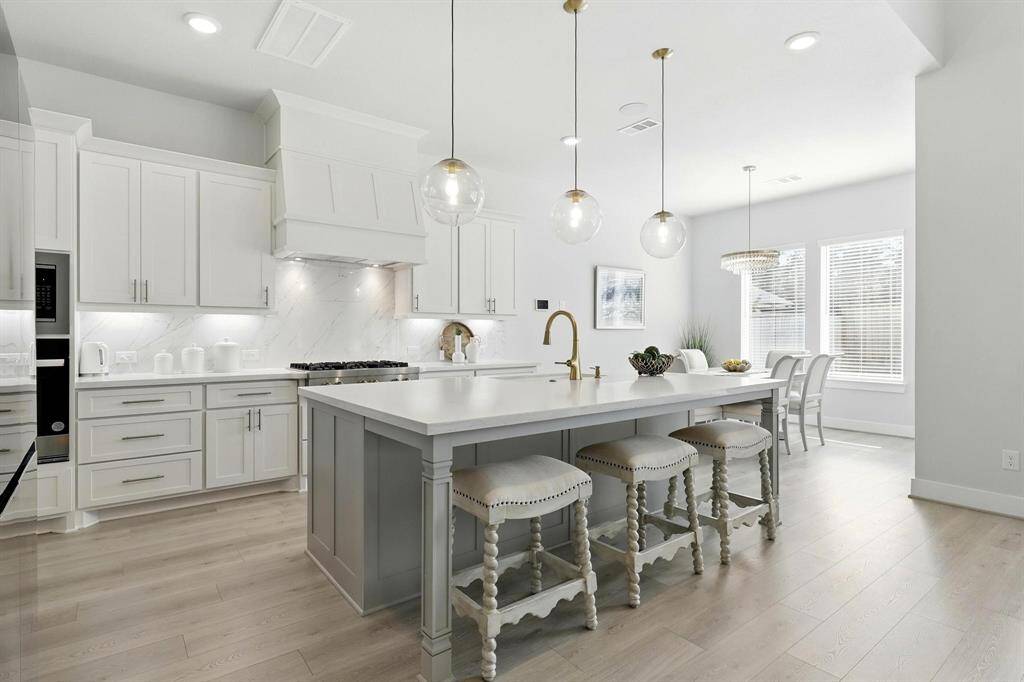 This kitchen features a sleek and modern design with white cabinetry, stainless steel appliances, and a spacious island with seating. Pendant lighting adds elegance, while an adjacent dining area offers ample space for entertaining. Light wood flooring enhances the airy, contemporary feel.