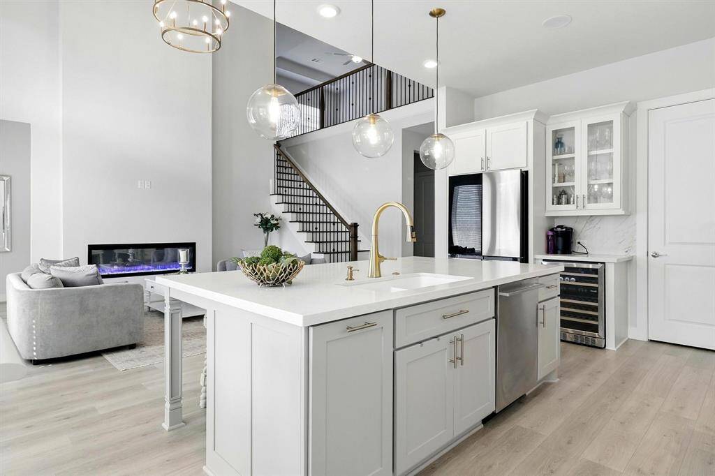 This modern kitchen features sleek white cabinetry, a spacious island with a gold-tone faucet, and pendant lighting. The open layout connects to a living area with a cozy fireplace and staircase, offering a bright and inviting space.
