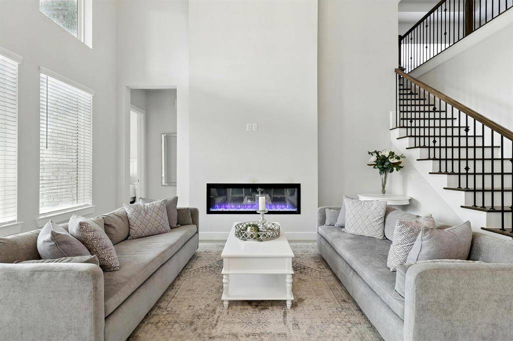 This inviting living room features high ceilings, large windows, and a modern electric fireplace.  and an open staircase with a sleek railing, creating a bright and spacious atmosphere.