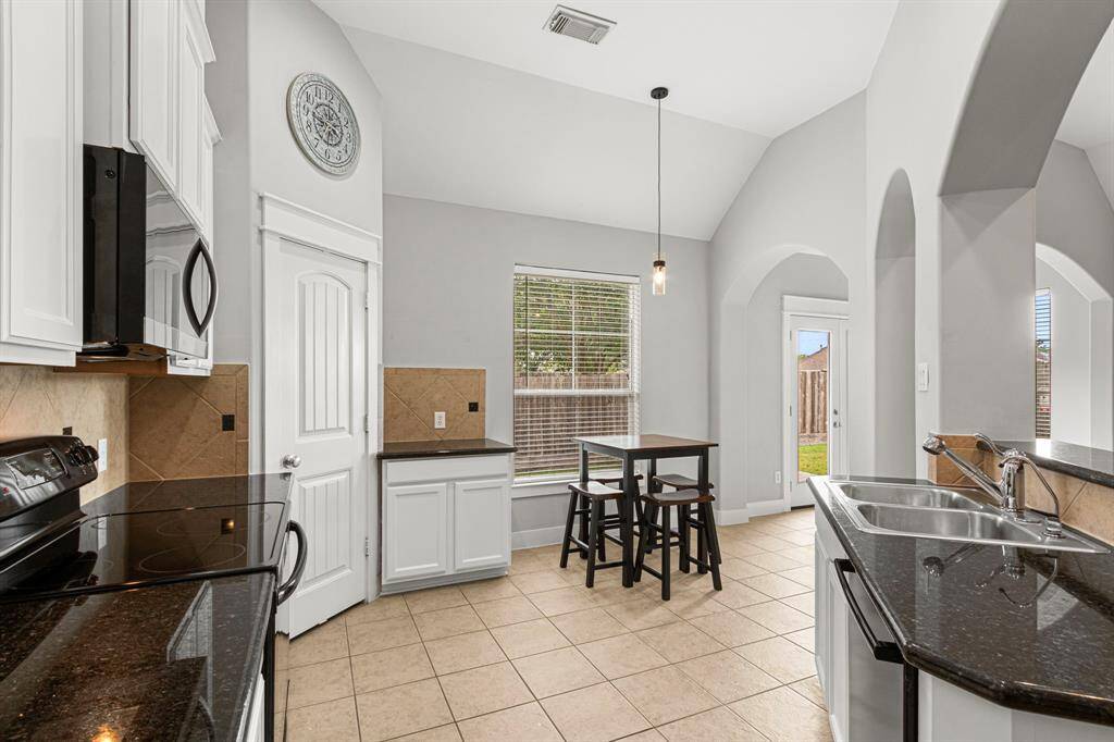 Bright kitchen with vaulted ceilings and modern lighting. Features black granite countertops, white cabinetry, and tiled flooring. Includes a cozy breakfast nook by a large window overlooking the backyard.