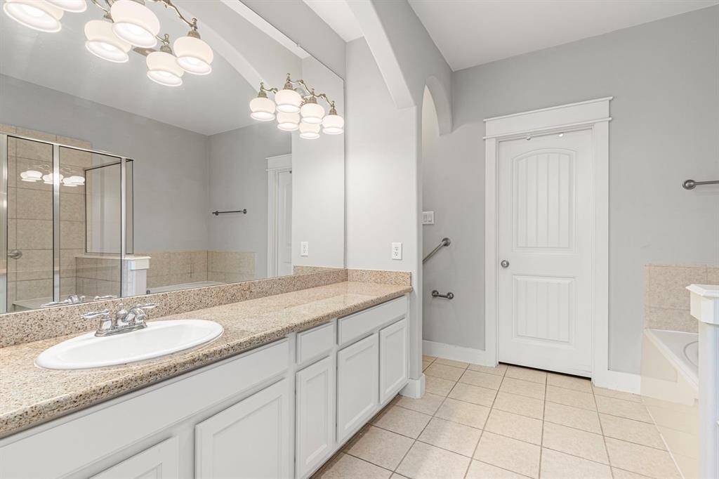 The primary bathroom features a spacious vanity with a granite countertop, ample lighting, and a large mirror. It includes a glass-enclosed shower, neutral tile flooring, and a separate bathtub, creating a bright and modern space.