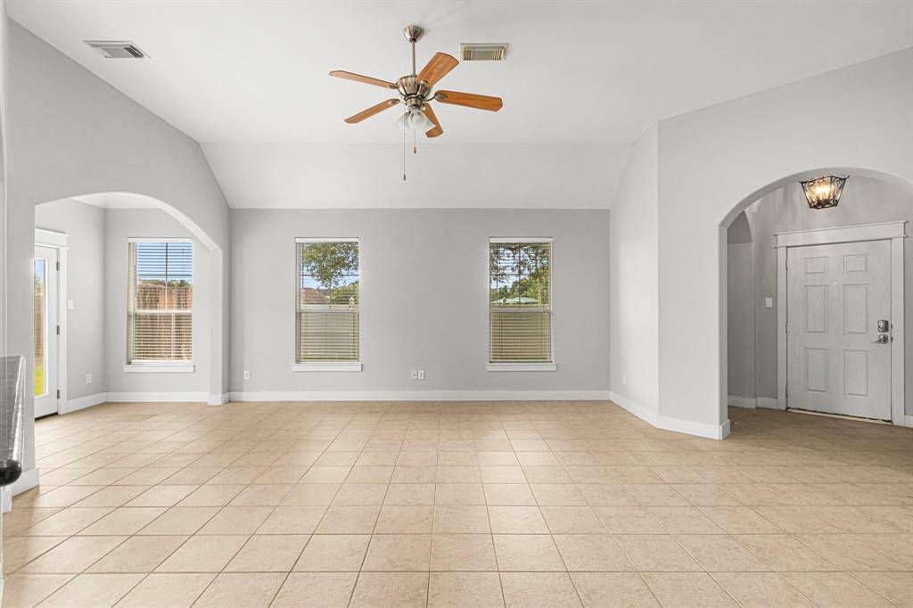 Bright and spacious living area with neutral tile flooring, high ceilings, and ample natural light from multiple windows. Features a ceiling fan, arched doorways, and a welcoming entryway. Ideal for comfortable living.