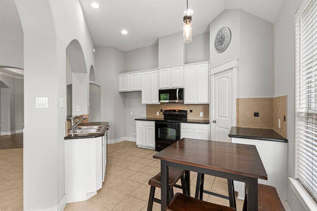 This kitchen features a modern design with high ceilings, white cabinetry, and black appliances. It includes a central island with a double sink and a casual dining area. The space is well-lit with natural light from large windows and has neutral tile flooring.