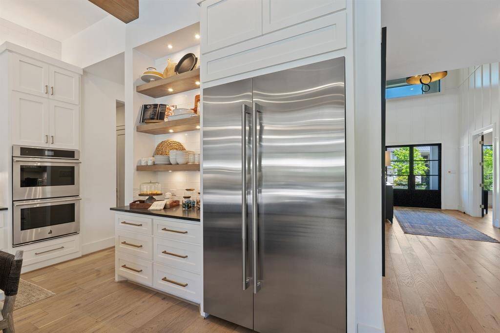 Sleek and sophisticated, this kitchen is equipped with all high end appliances and this section of the kitchen features a built-in stainless steel refrigerator framed by custom cabinetry and soft-close drawers. Open shelving with accent lighting adds both style and function, perfect for showcasing dishes or serving essentials. The view also highlights the beautiful flow into the grand foyer, with wide-plank wood flooring and natural light pouring through oversized windows.