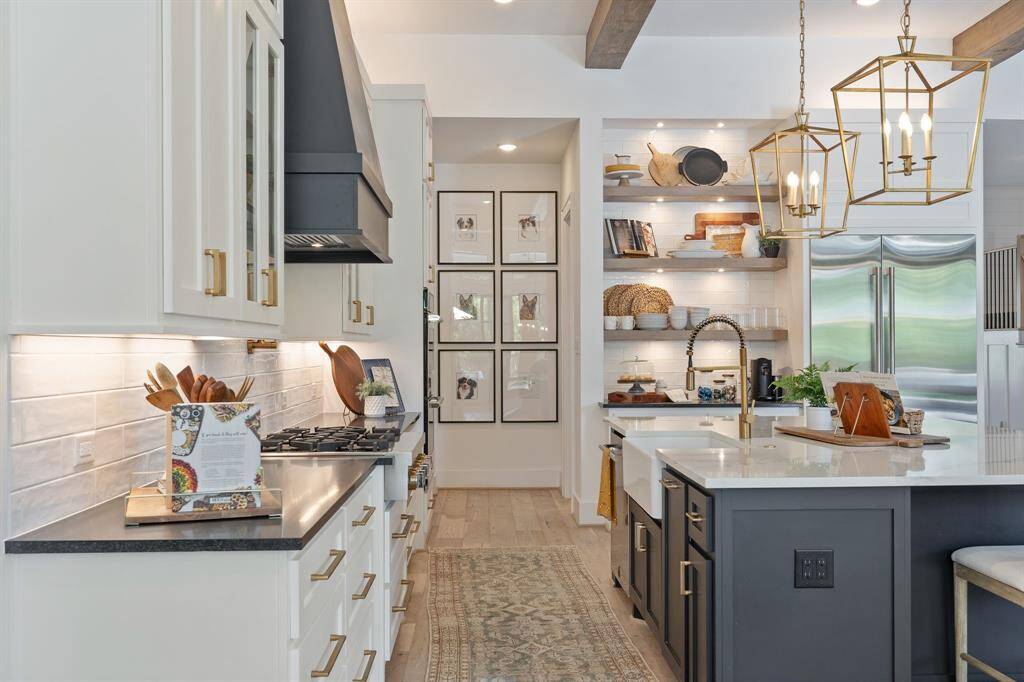 This image showcases a stunning view of the heart of the kitchen, blending elegance and function. On the left, sleek honed granite meet crisp white cabinetry with elegant hardware and under-cabinet lighting. A striking vent hood adds bold contrast above the gas cooktop. To the right, the expansive island features a deep farmhouse sink and plenty of seating, all under statement-making gold lantern pendant lights.