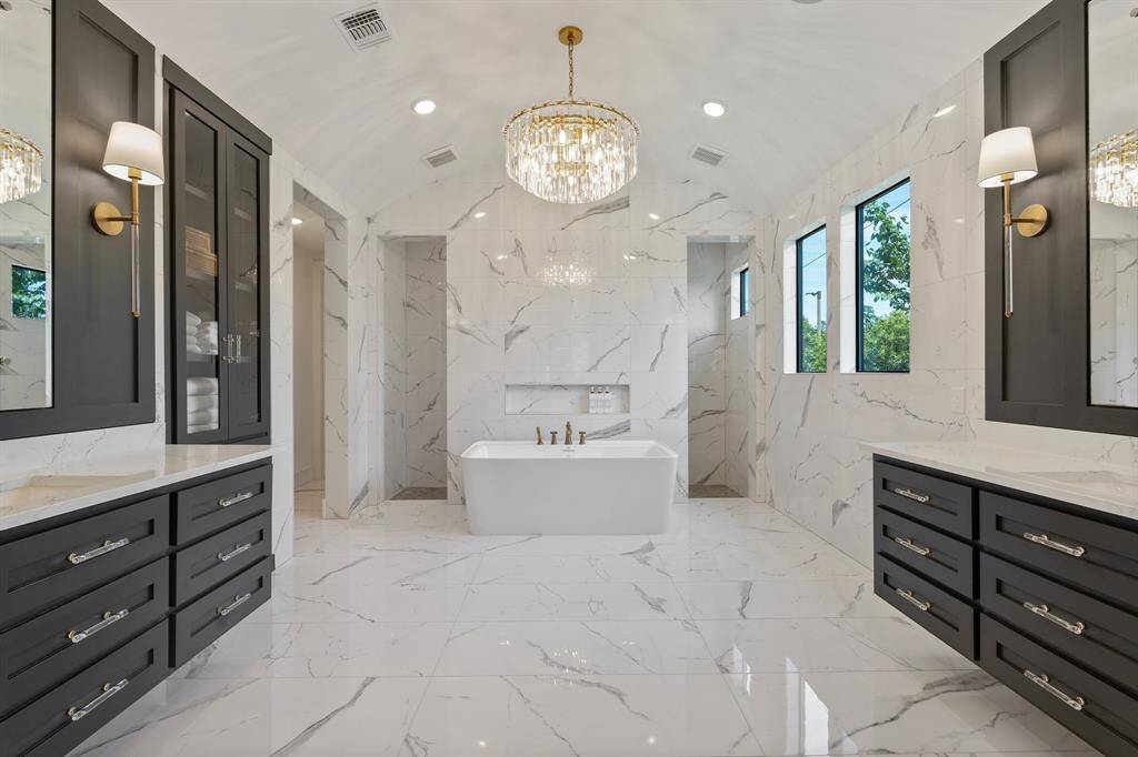 Luxurious spa-inspired bathroom featuring dramatic barrel ceiling, gleaming marble-look tile throughout, dual vanities, and a statement chandelier above a freestanding soaking tub. Beyond the tub is the dual walk in shower.
