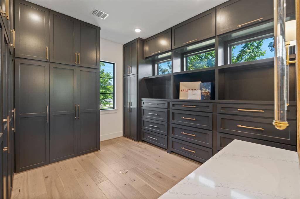 A dream-worthy closet with extensive built-ins, soft-close cabinetry, natural light, and luxurious hardware—designed for both beauty and functionality. To the right, a spacious vanity with a 6' mirror and elegant sconces offers a glamorous and convenient space to get ready.