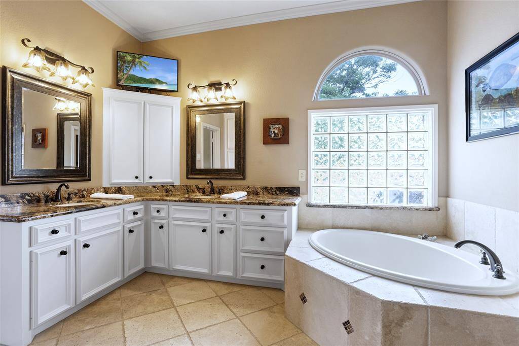 The ensuite Primary Bathroom offers a spacious layout with dual sinks, elegant granite countertops, and ample built-in cabinetry. A large soaking tub is set against a glass block window, allowing natural light to flood the room and creating a relaxing and serene atmosphere.