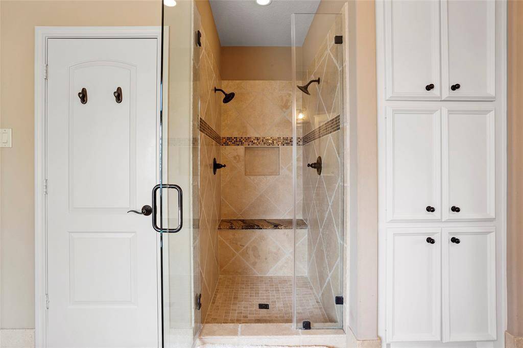 The modern, tiled walk-in shower is equipped with dual showerheads, bench seating, and glass doors, creating a spa-like experience. It is conveniently adjacent to built-in white storage cabinets, combining sleek design with functional storage in the Primary Bathroom.