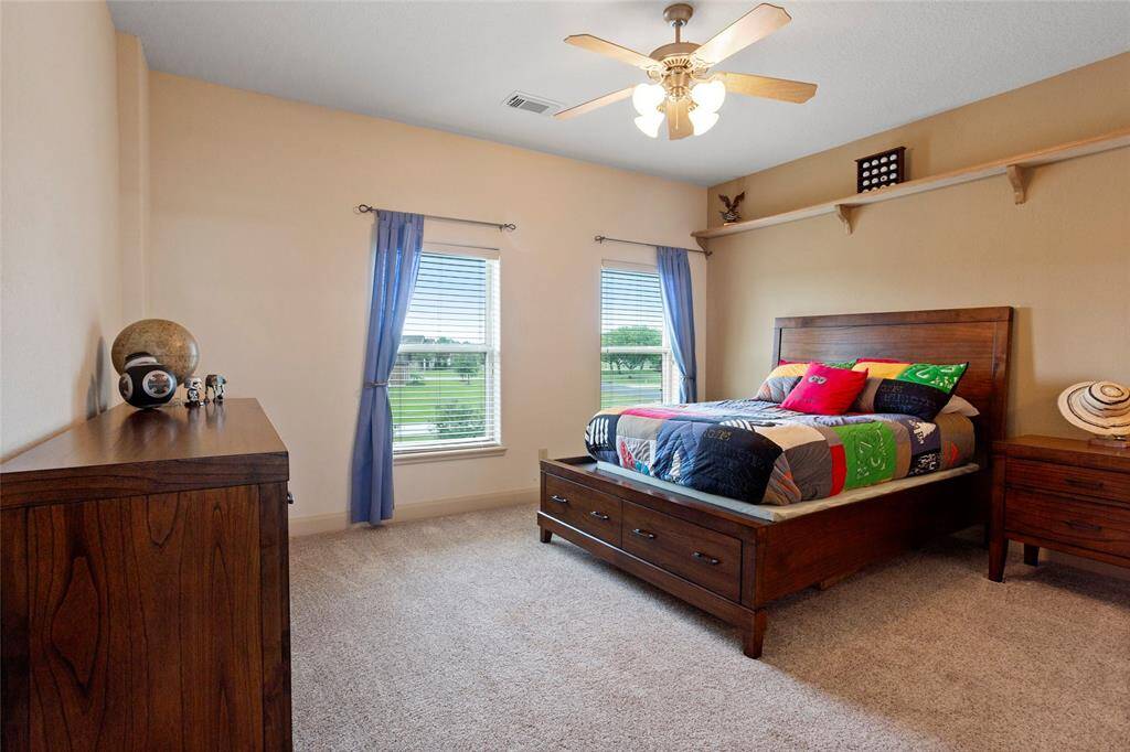 This upstairs bedroom features a large walk-in closet with built-in shelving, neutral walls, carpeted flooring, and an ensuite Hollywood bathroom. Two windows fill the room with natural light, while a shelf above the bed provides extra storage or display space.