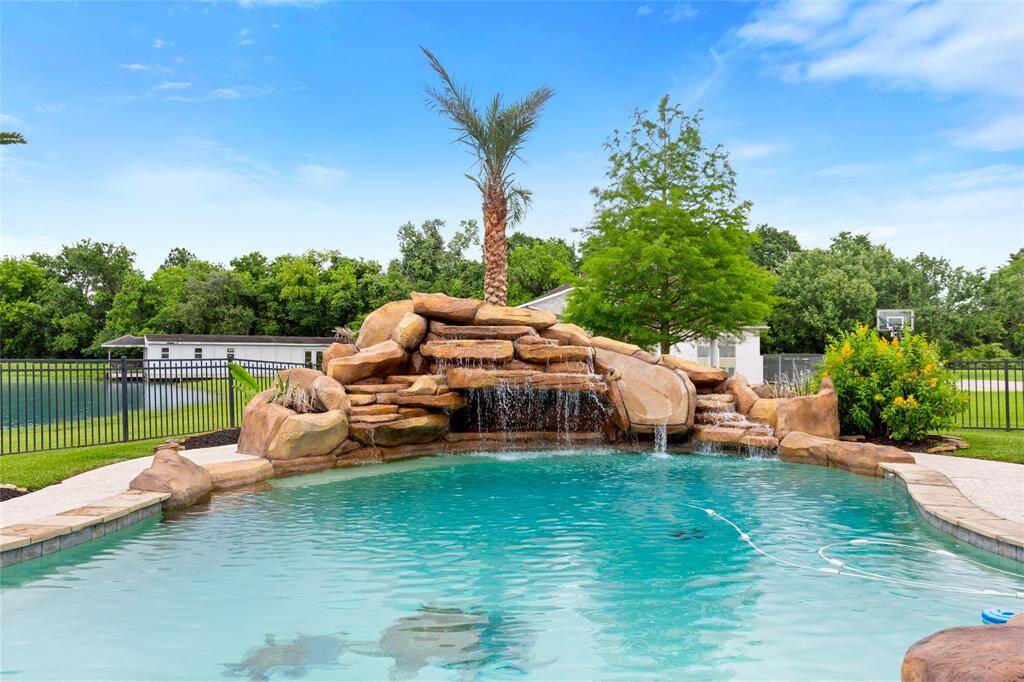 This photo features a The beautifully landscaped backyard showcases a large pool accented by a stunning rock waterfall, slide, and grotto feature. Surrounded by lush greenery, the space extends to the lake just beyond the iron fence, creating a tranquil outdoor oasis ideal for both relaxation and entertaining.