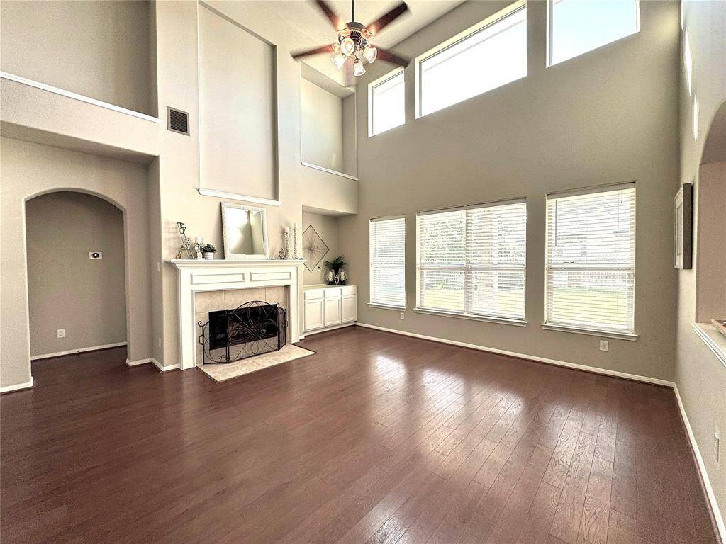 This spacious family room features high ceilings, abundant natural light from the wall of windows, and elegant wood flooring. A cozy fireplace adds charm, and the neutral color palette offers a modern and inviting atmosphere.