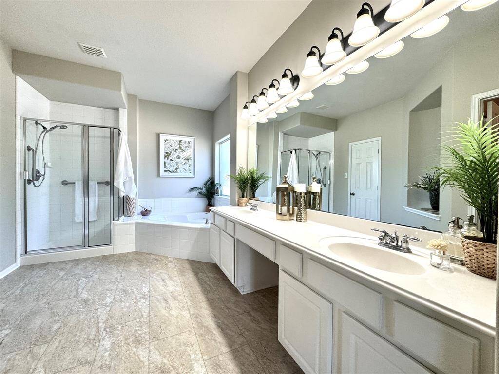 Primary bathroom features a large dual-sink vanity with ample lighting, a glass-enclosed shower, and a soaking tub. The neutral tile flooring and soft color palette create a calming atmosphere, complemented by elegant fixtures and art niche.