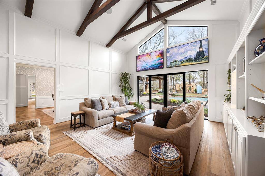 Living room features neutral tones, high ceilings with wooden beams, large, black-framed windows that provide ample natural light, and a cozy seating area. The room is accented with built-in shelves and overlooks a well-maintained outdoor area.
