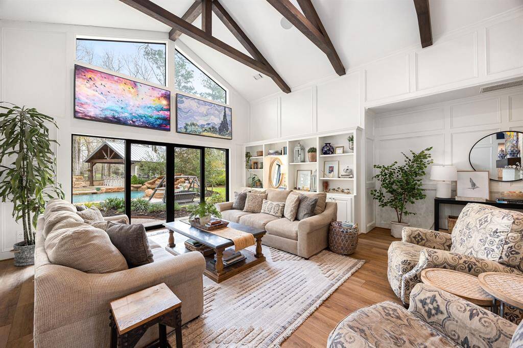 This spacious living room features a high vaulted ceiling with exposed wooden beams, built-in shelving, and large windows offering abundant natural light and views of the backyard. Custom placed tv monitors fit perfectly over the windows. Additional storage closet hidden in the wall.