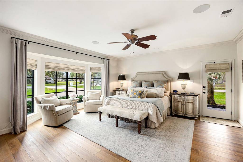 The spacious primary retreat features a cozy seating area surrounded by large windows, allowing plenty of natural light. It has elegant decor with neutral tones, a ceiling fan, recessed speakers, built-in shelving and cabinetry and a door leading outside to a large covered side porch.