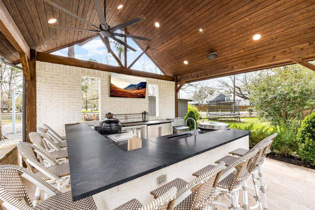 This outdoor space features a spacious, covered patio with a modern kitchen and bar area, equipped with a sleek countertop, ample seating, and built-in appliances. The area is surrounded by lush greenery, offering a perfect setting for entertaining and relaxation.