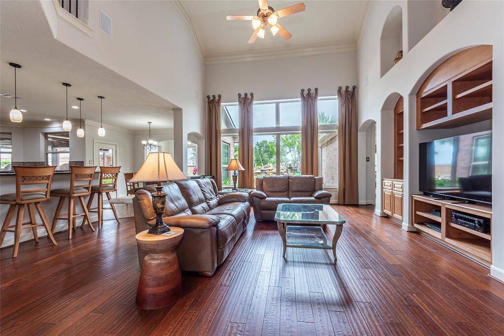 The spacious family room features fresh neutral paint tones, soaring ceiling, and floor-to-ceiling windows that flood the space with natural light. Warm wood flooring enhances the inviting atmosphere, while the open-concept design seamlessly connects to the kitchen, breakfast area, and sunroom, creating the perfect setting for everyday living.