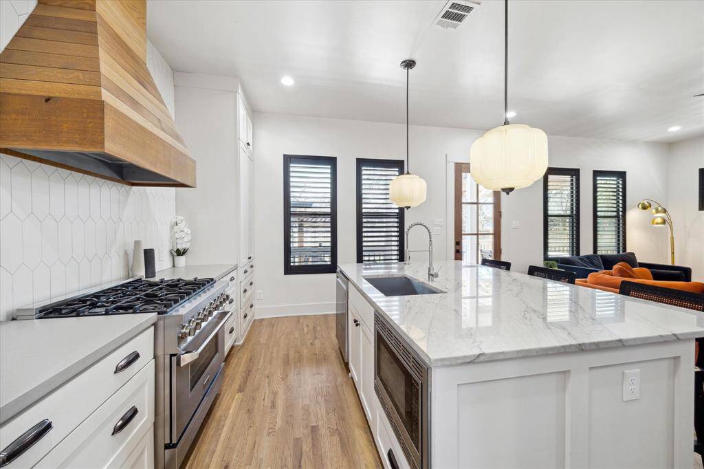 Modern kitchen filled with designer touches. Crisp white custom cabinetry stretches to the ceiling, accented by sleek black hardware and glass-paneled upper cabinets. A striking wooden range hood brings warmth and contrast against the clean, textured backsplash.