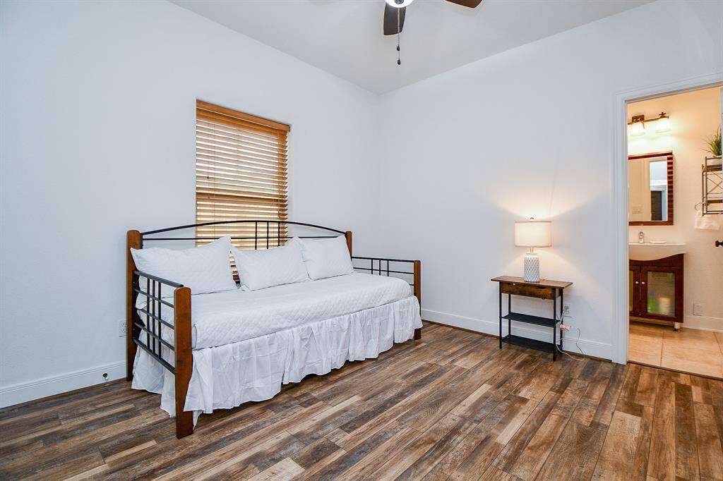 This cozy bedroom features wood-style flooring, a daybed with white bedding, and a bedside table with a lamp. It includes a window with blinds and an en suite bathroom for added convenience.