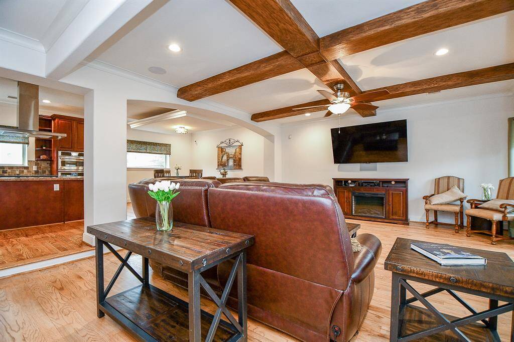 This spacious living room features hardwood floors, exposed wooden beams, and a ceiling fan. It includes a cozy seating area with leather couches, a mounted TV, and a fireplace. The open layout connects to a modern kitchen with stainless steel appliances and a dining area, creating an inviting atmosphere for family gatherings.