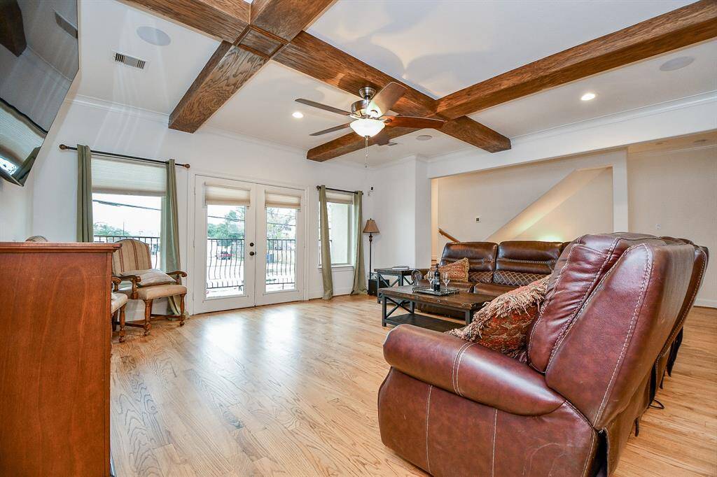 This spacious living room features hardwood floors, a ceiling fan, and exposed wooden beams for a cozy, rustic feel. Large windows and French doors provide ample natural light, leading to a balcony. The room is furnished with comfortable leather seating and a coffee table.