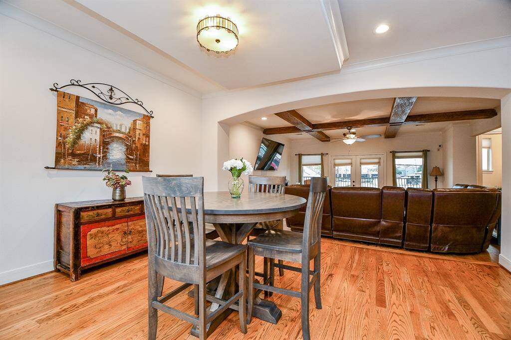 This inviting living area features an open floor plan with elegant wood flooring, a cozy dining space, and a comfortable seating area. The room is accentuated by rustic ceiling beams and ample natural light from large windows and glass doors, enhancing its warm and welcoming atmosphere.