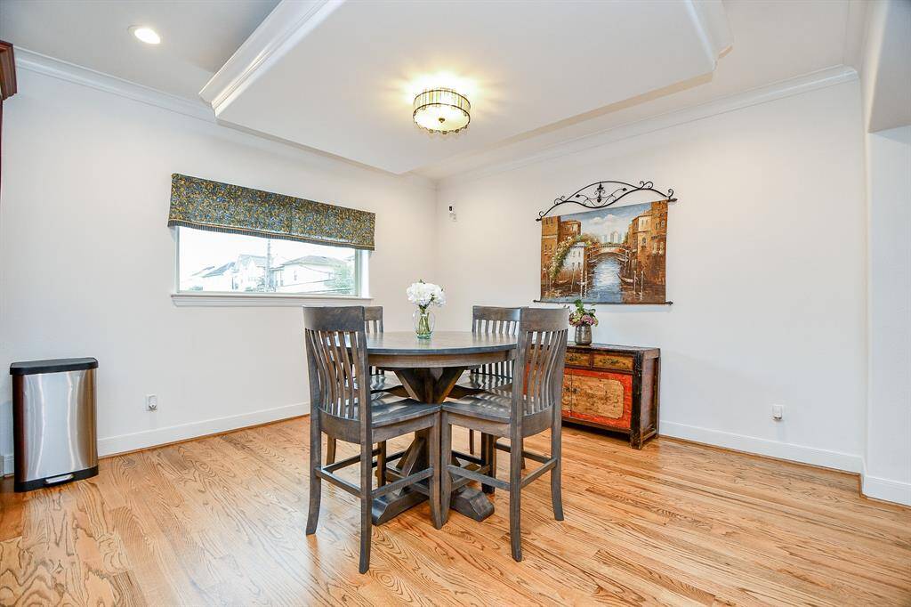 This dining area features hardwood floors, a stylish light fixture, and ample natural light from a window with a patterned valance. The space includes a chic round table with four chairs and a decorative sideboard with artwork above, creating a cozy and inviting atmosphere.