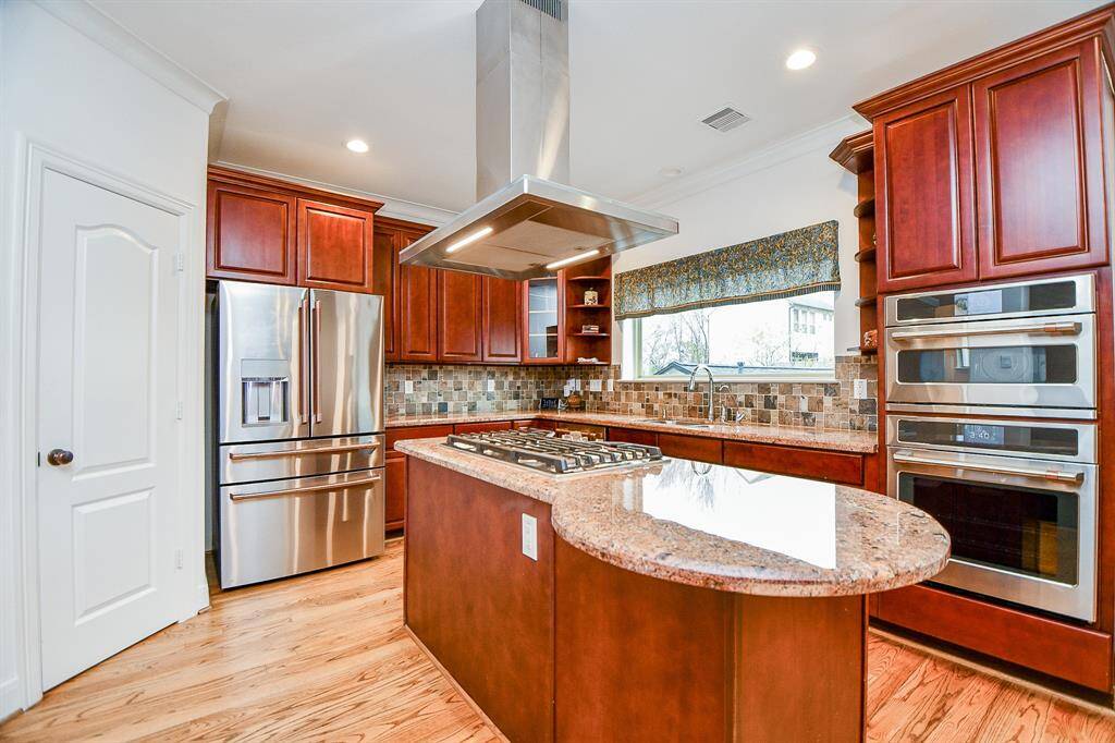 This kitchen features elegant wooden cabinetry, stainless steel appliances, a central island with a built-in gas cooktop, and a stylish tile backsplash. The natural light from a large window enhances the warm tones, and the double wall ovens add modern convenience.