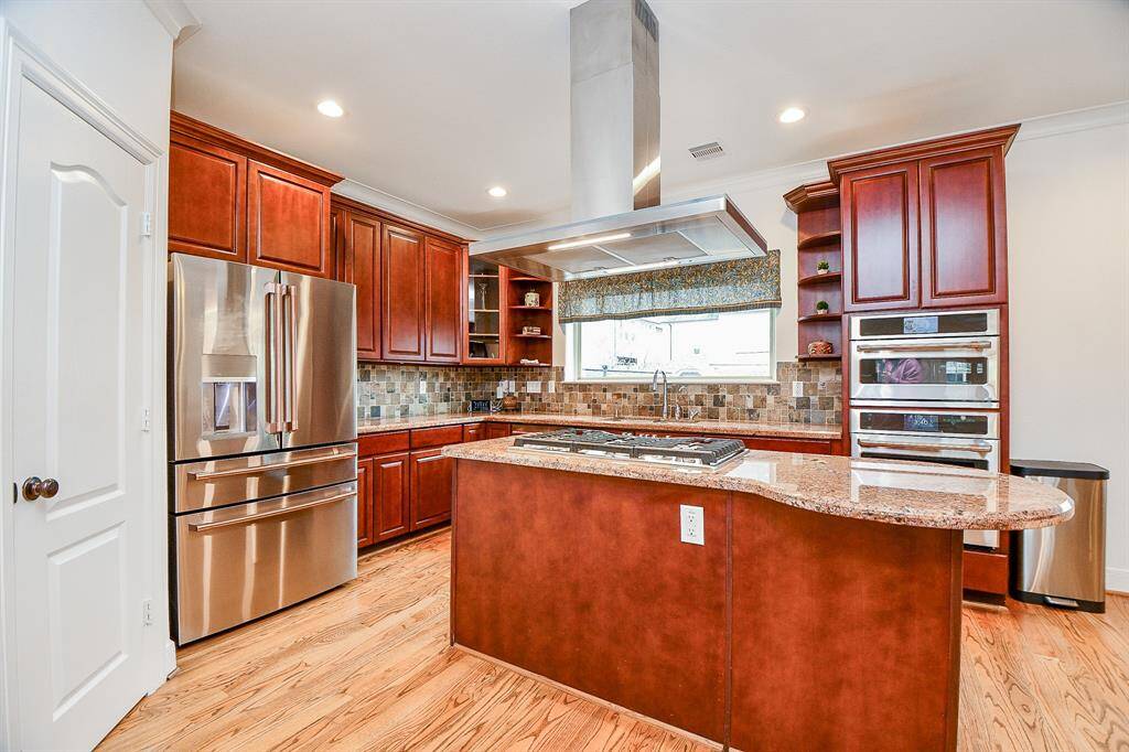 This kitchen features rich wooden cabinetry, stainless steel appliances, and a central island with a gas cooktop. The space is well-lit with recessed lighting and has a stylish tile backsplash, creating a warm and inviting atmosphere.