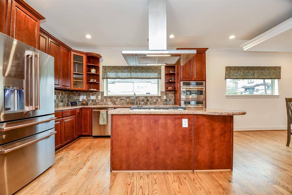 This modern kitchen features rich wood cabinetry, stainless steel appliances, and a central island with a gas cooktop and overhead range hood. It offers ample counter space, a tiled backsplash, and natural light from two windows, creating a warm and functional cooking environment.