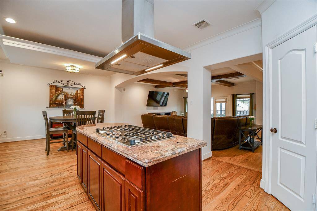 This photo showcases an open-concept living area with warm hardwood floors. It includes a kitchen island with a cooktop, a dining area with a table, and a cozy living room with a mounted TV. The space is well-lit with a modern ceiling light and features stylish coffered ceilings.