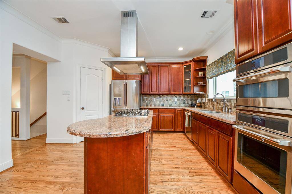 This kitchen features rich wooden cabinetry, granite countertops, and stainless steel appliances. It includes a central island with a cooktop and a modern range hood. The space is brightened by a large window and wood flooring, offering a warm and inviting atmosphere.