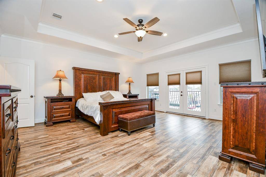 This spacious bedroom features elegant wood flooring, a ceiling fan, and abundant natural light from large windows. It includes a stylish wooden bed, matching nightstands, and a cozy bench, creating a warm, inviting atmosphere.